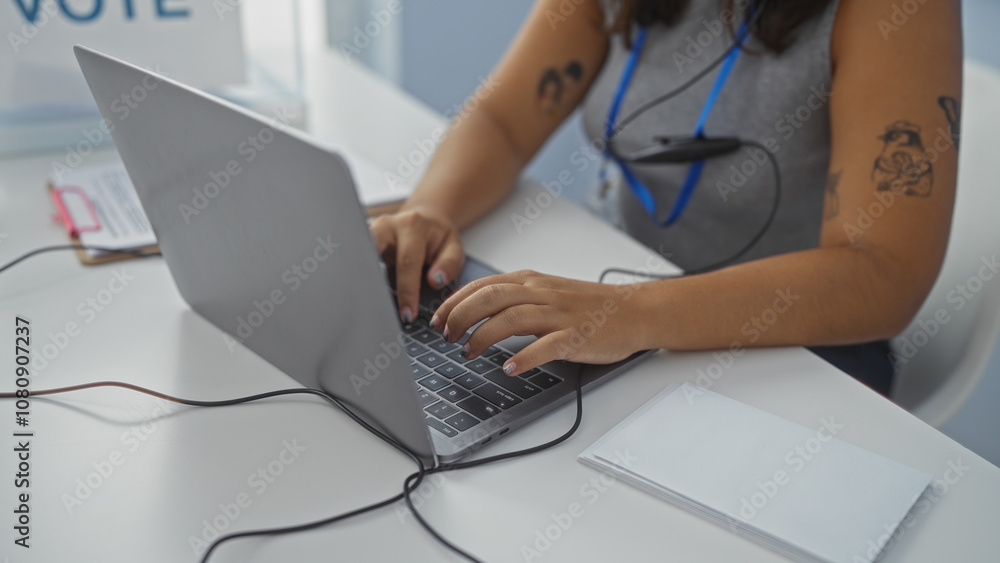 Young hispanic woman in an indoor electoral college setting typing on a laptop, with documents visible on table. Young hispanic woman in an indoor electoral college setting typing on a laptop, with documents visible on table.