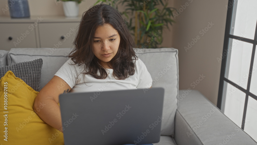 A young hispanic brunette woman is working on a laptop in a cozy living room setting, with a grey sofa and indoor plants in the background, embodying a relaxed and focused atmosphere. A young hispanic brunette woman is working on a laptop in a cozy living room setting, with a grey sofa and indoor plants in the background, embodying a relaxed and focused atmosphere.