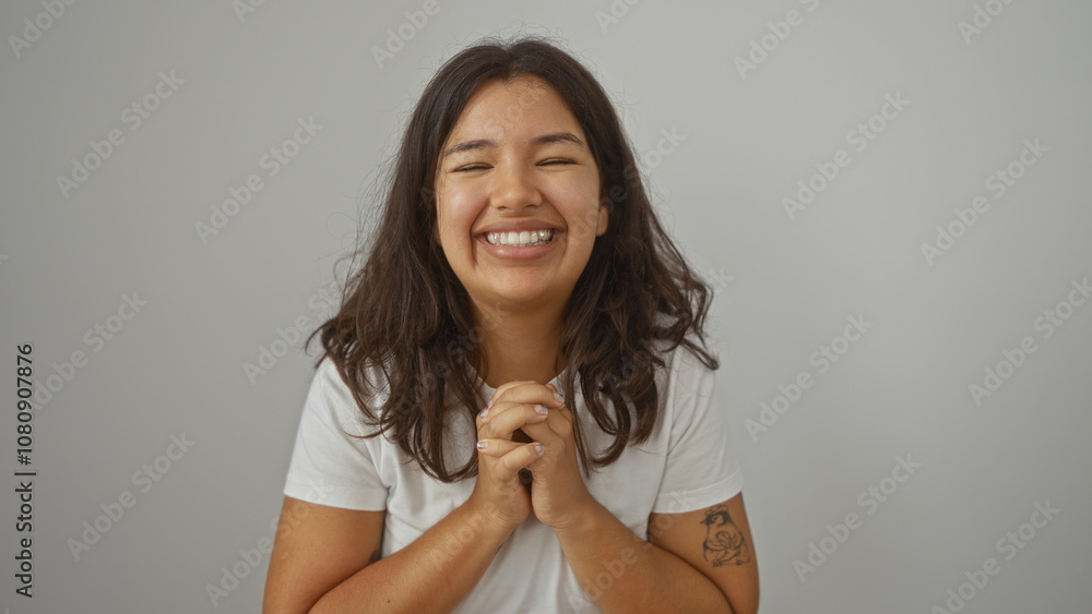 A beautiful young hispanic woman with brunette hair smiling joyfully against a white wall, wearing a casual white shirt and displaying a tattoo on her arm. A beautiful young hispanic woman with brunette hair smiling joyfully against a white wall, wearing a casual white shirt and displaying a tattoo on her arm.