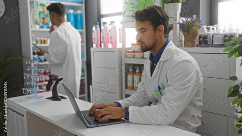 Two hispanic male pharmacists working together in a pharmacy with one using a laptop and the other organizing shelves indoors.