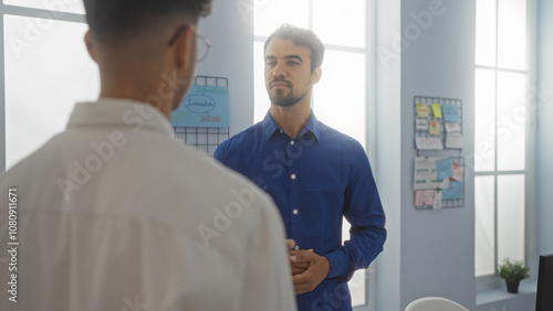 Two hispanic men, workers in a business setting, stand together in an indoor workplace room with bright windows and office materials in the background.