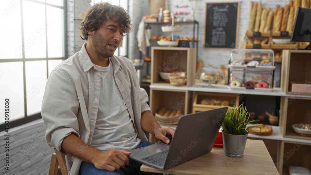 Young man working on laptop in modern bakery with shelves of bread and pastries in background Young man working on laptop in modern bakery with shelves of bread and pastries in background