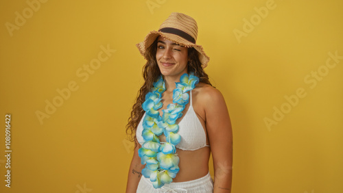 Young woman winking over isolated yellow background wearing a hat and floral lei