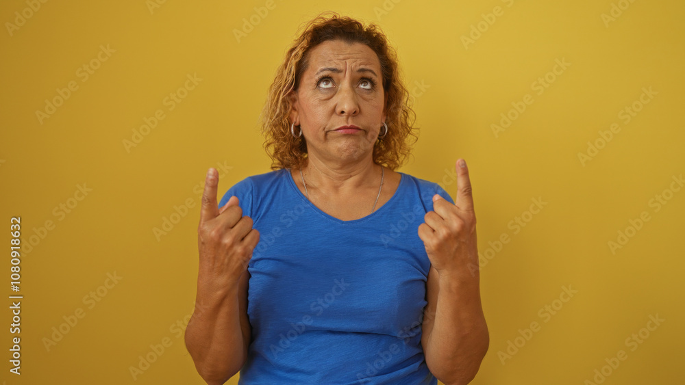 Middle-aged hispanic woman with curly hair in a blue shirt standing against a yellow wall, pointing upward and looking upwards. Middle-aged hispanic woman with curly hair in a blue shirt standing against a yellow wall, pointing upward and looking upwards.