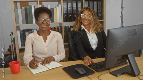 Women working together in an office, with one writing in a notebook and the other working on a computer, both smiling in a professional indoor workplace setting. photo