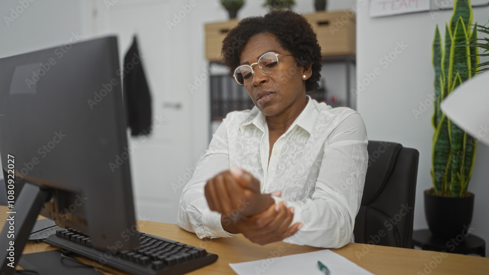 Woman working on a computer in an office while holding her wrist, indicating discomfort, with curly hair and glasses, surrounded by indoor plants and office decor in the background Woman working on a computer in an office while holding her wrist, indicating discomfort, with curly hair and glasses, surrounded by indoor plants and office decor in the background