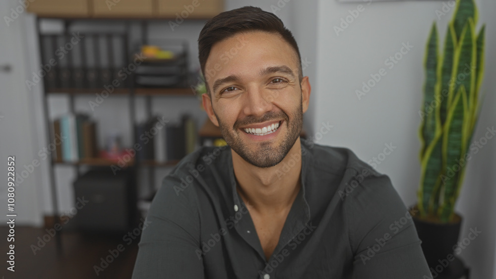 Handsome young hispanic man with a beard smiling in an indoor office setting with shelves and a plant in the background. Handsome young hispanic man with a beard smiling in an indoor office setting with shelves and a plant in the background.
