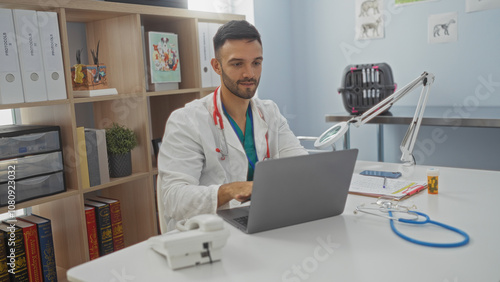 Young handsome hispanic male veterinarian working indoors at a clinic on his laptop, surrounded by medical equipment, shelves with books, and files.