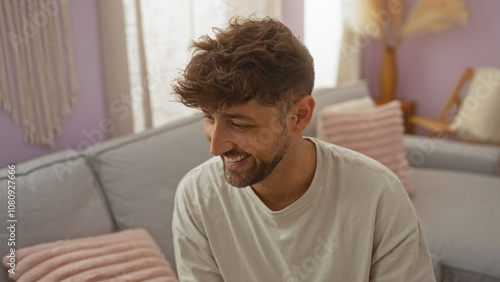 Young man smiling indoors in a cozy living room setting with pastel decor and plush cushions, showcasing a relaxed home atmosphere