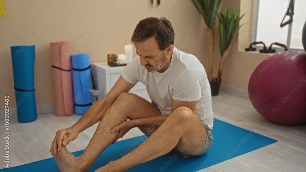 Hispanic man sitting on mat at wellness center massaging sore leg surrounded by yoga equipment and candlelight in a peaceful indoor setting. Hispanic man sitting on mat at wellness center massaging sore leg surrounded by yoga equipment and candlelight in a peaceful indoor setting.