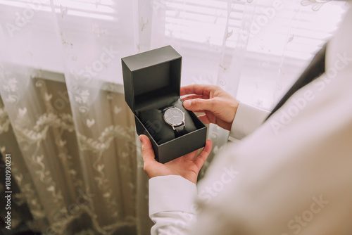 A man is holding a Rolex watch in a black box. The watch is a luxury brand and the box is made of high-quality materials. The man is wearing a white shirt and a black tie