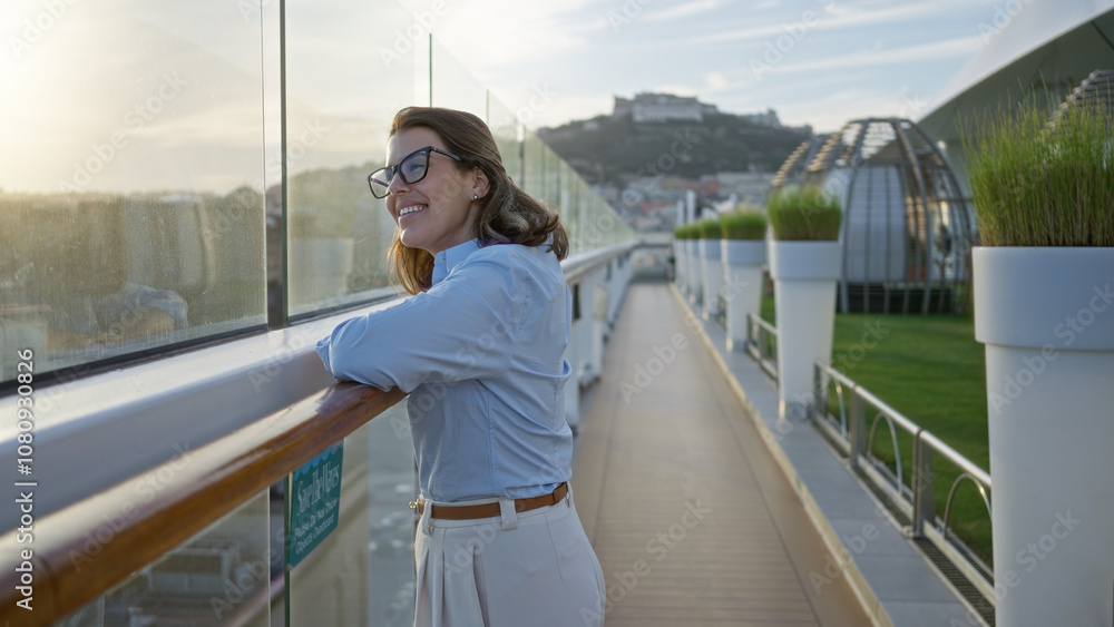 Obraz premium Woman smiling on cruise ship outdoors at dock enjoying scenery in the evening light with city view in the background and white planters lining the deck