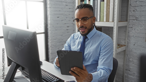 Young man with braids using tablet in modern office setting, wearing glasses and blue shirt, surrounded by contemporary interior and technology elements, showcasing focus and engagement.