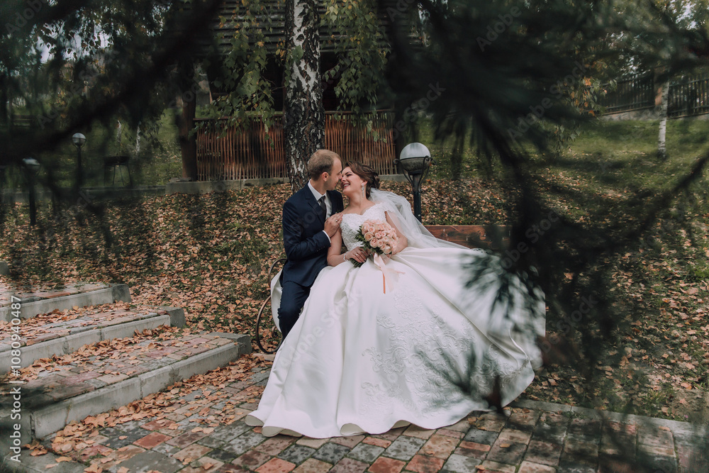 A bride and groom are sitting on a bench in a park. The bride is wearing a white dress and the groom is wearing a suit. They are holding hands and smiling at the camera A bride and groom are sitting on a bench in a park. The bride is wearing a white dress and the groom is wearing a suit. They are holding hands and smiling at the camera