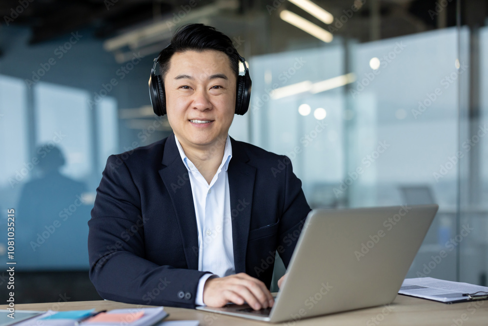 Portrait of a confident and confident Asian man in a suit and headphones sitting in the office at a laptop and looking at the camera with a smile Portrait of a confident and confident Asian man in a suit and headphones sitting in the office at a laptop and looking at the camera with a smile