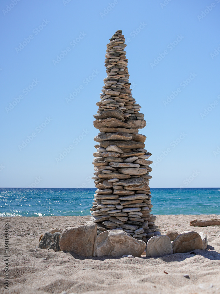 A tall, intricate stack of flat stones standing on a sandy beach against the backdrop of a calm blue sea and clear sky, symbolizing balance, meditation, and patience. A tall, intricate stack of flat stones standing on a sandy beach against the backdrop of a calm blue sea and clear sky, symbolizing balance, meditation, and patience.