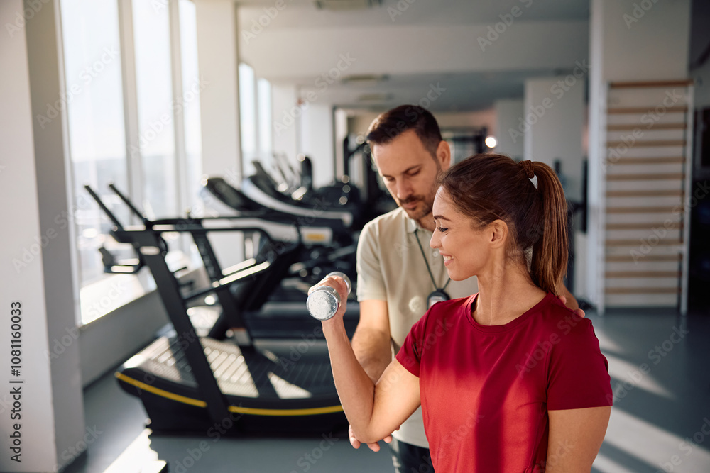 Happy woman exercising with hand weighs during sports training with her instructor in a gym. Happy woman exercising with hand weighs during sports training with her instructor in a gym.
