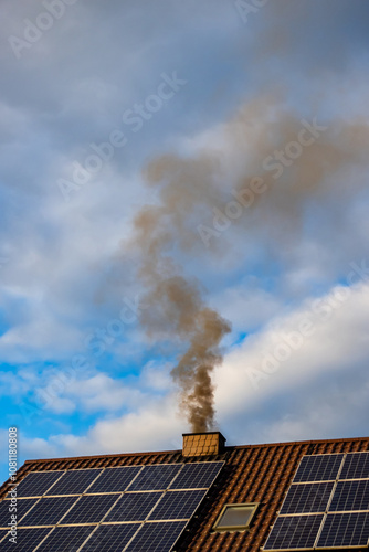Obraz na płótnie Black smoke from the chimney of a single-family home. Roof covered with solar panels. Roof against a blue, slightly cloudy sky. w salonie