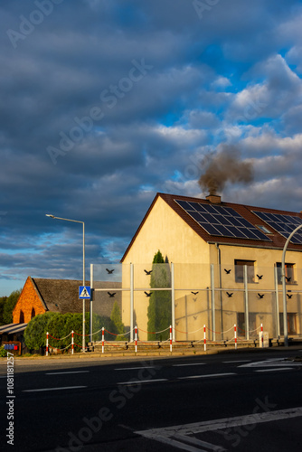 Black smoke from the chimney of a single-family home. Roof covered with solar panels. Roof against a blue, slightly cloudy sky.