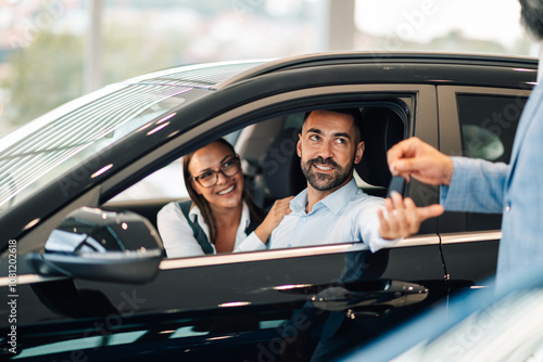 Happy couple inside car receiving keys from a salesperson in a dealership