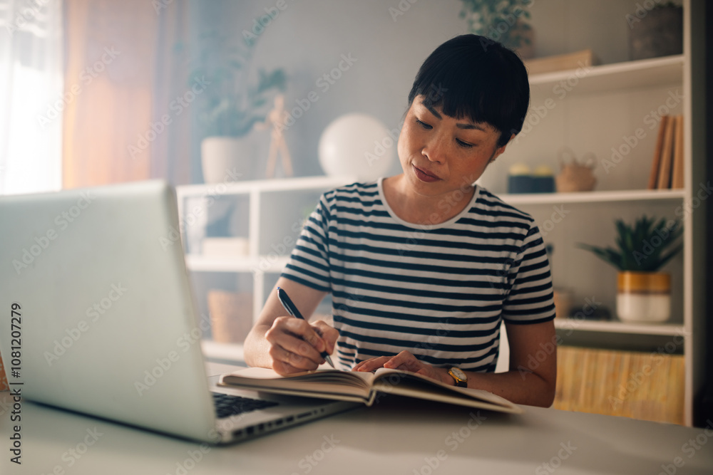 Focused asian woman writing in a notebook with a laptop on the desk. Focused asian woman writing in a notebook with a laptop on the desk.