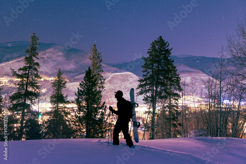 Hiker with snowboard on their back walking through the snow above a illuminated mountain town below
