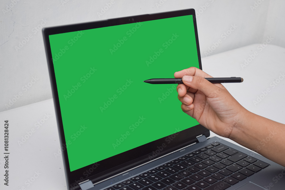Close-up of a hand holding a stylus, pointing at a blank green screen on a laptop. set up for digital design or illustration Close-up of a hand holding a stylus, pointing at a blank green screen on a laptop. set up for digital design or illustration
