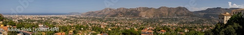 Panoramic view of valley dotted with houses, framed by towering mountains and historic buildings perched on hill