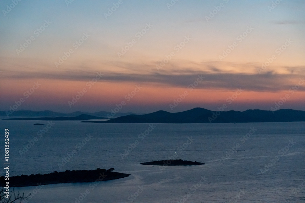 Obraz premium Serene sunset view over tranquil sea and silhouette of islands with vibrant sky colors, showcasing peaceful twilight and natural beauty.
