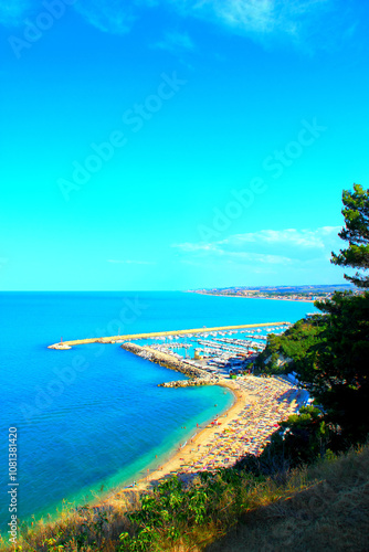 Dreamy scene from Numana Alta with the port of Numana and its many docked white boats surrounded by rocks washed by the Adriatic Sea, thriving green vegetation, sandy beach with resorts and equipment