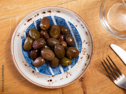 On small square white plate lies portion of large ripe green olives with stone. Snack is supplemented with glass of beer photo