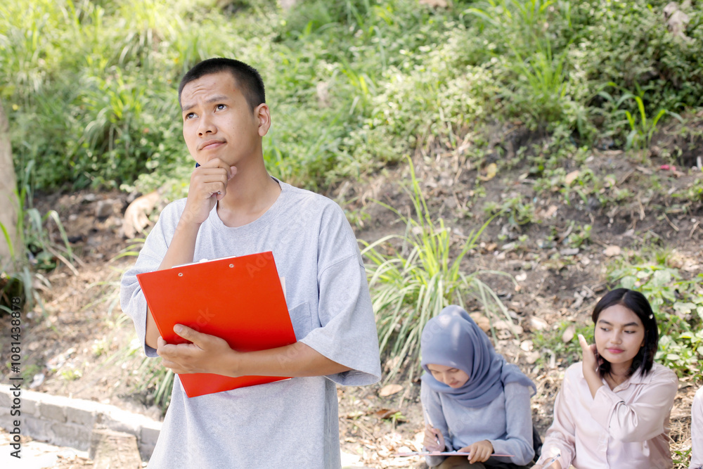 Asian Man Student Holds A Clipboard Showing Thinking Gesture In Outdoor Learning Activity With Friends Asian Man Student Holds A Clipboard Showing Thinking Gesture In Outdoor Learning Activity With Friends