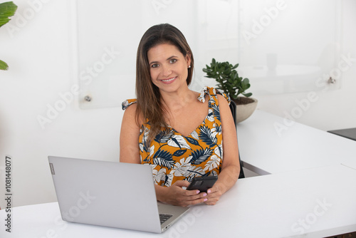 Smiling woman sitting at desk with laptop and smartphone in modern office, wearing a floral dress, represents workplace productivity, communication, career success and confident business style