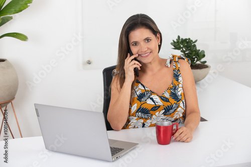 Confident woman using smartphone while seated at desk with laptop and red coffee mug, wearing floral blouse in a modern office setting, showcasing multitasking, leadership, and workplace efficiency