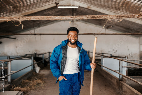 Portrait of a black smiling barn worker posing with pitchfork in stable with cows.