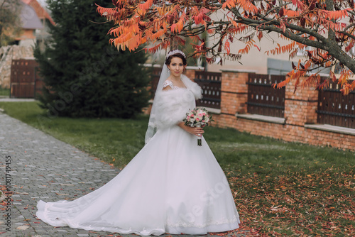 A bride is standing in front of a tree with a white dress and a fur stole. She is holding a bouquet of flowers photo