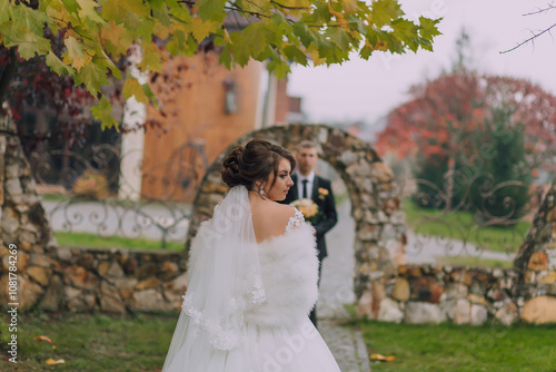 A bride and groom are standing in front of a stone archway. The bride is wearing a white dress and a fur stole, while the groom is wearing a suit. The scene is set in a garden photo