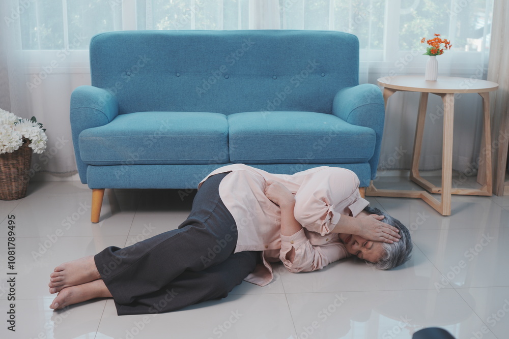 Unhappy Asian senior woman suffering from severe stomach pain lying on the floor in front of the living room sofa at home