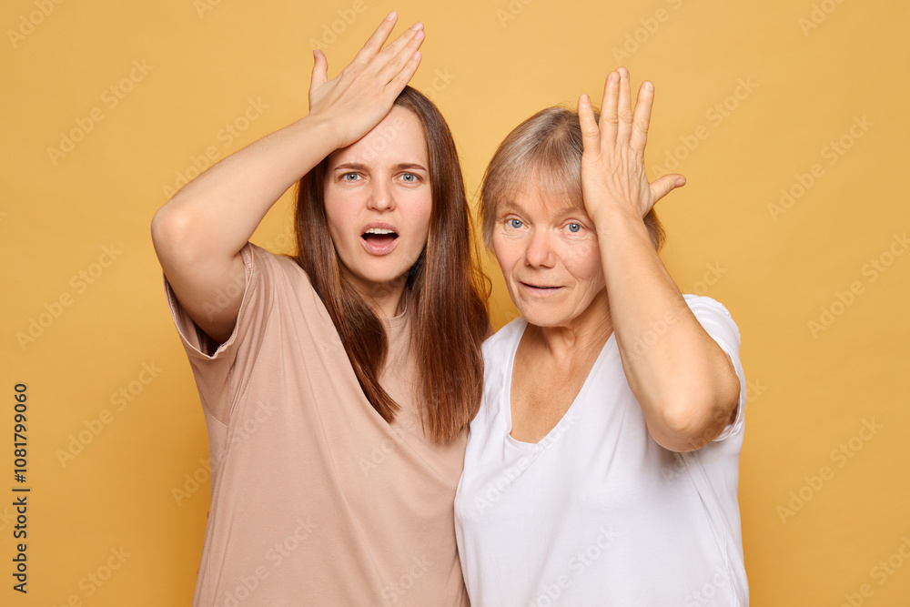 Two women displaying surprise and disbelief against a vibrant yellow background while touching their foreheads in a playful manner, showcasing their expressions during a lighthearted moment Two women displaying surprise and disbelief against a vibrant yellow background while touching their foreheads in a playful manner, showcasing their expressions during a lighthearted moment