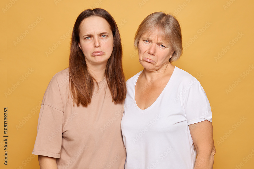Two women with serious expressions pose against a yellow background in a casual setting, showcasing contrasting emotions while standing side by side Two women with serious expressions pose against a yellow background in a casual setting, showcasing contrasting emotions while standing side by side