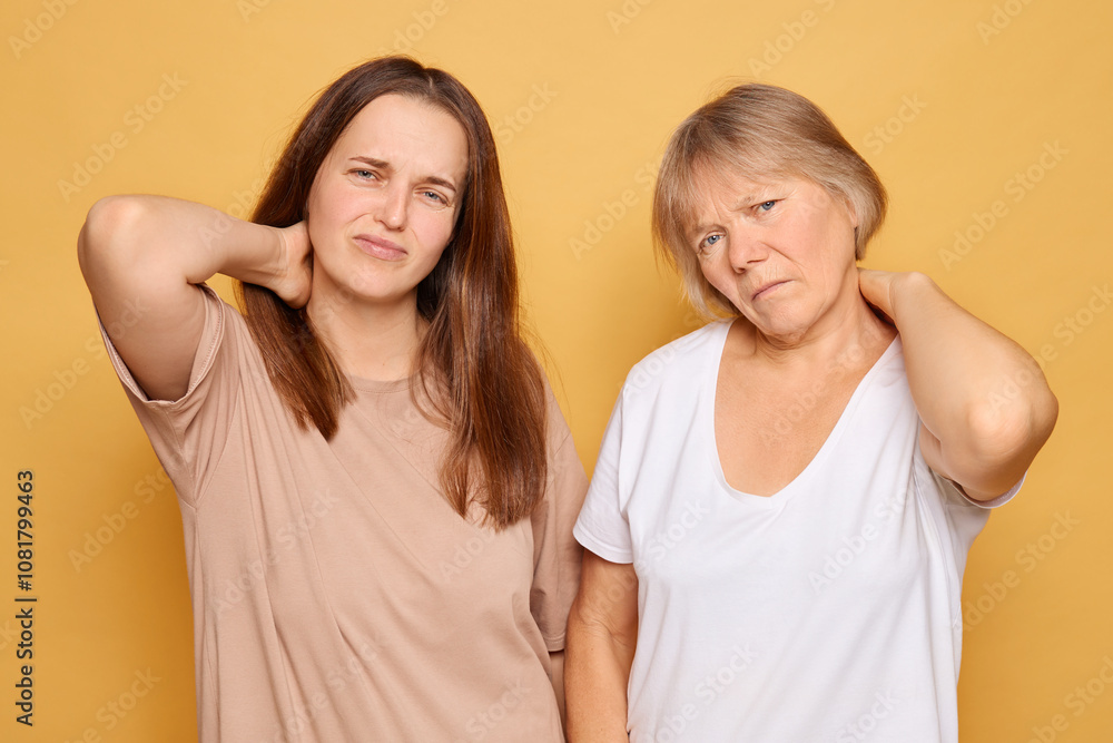 Two women standing against a yellow background, both displaying expressions of discomfort while touching their necks in a casual indoor setting Two women standing against a yellow background, both displaying expressions of discomfort while touching their necks in a casual indoor setting