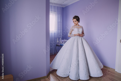 A woman in a white dress stands in front of a purple wall. She is wearing a long white gown and is posing for a picture