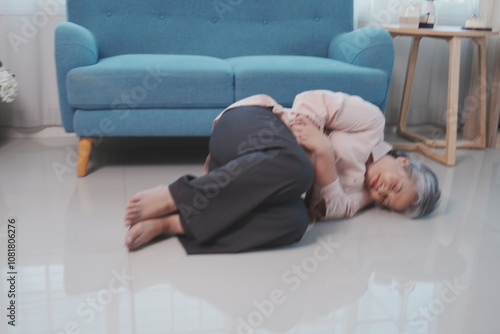 An elderly woman with gray hair falls down on the floor of a room with shoulder pain, suffering from pain in her stomach in the living room at home.