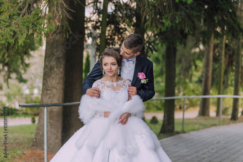 A bride and groom are posing for a picture in a park. The bride is wearing a white dress and a fur stole, while the groom is wearing a suit. Scene is happy and romantic photo