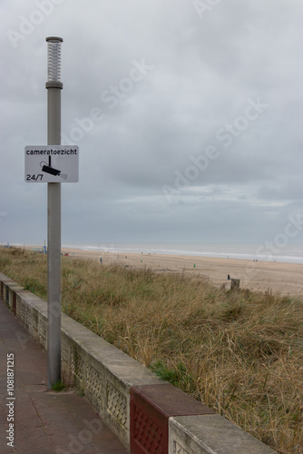 Coastal Path with 'Cameratoezicht 24/7' Surveillance Sign on Overcast Day: Beachside Walkway Monitored for Safety Under Cloudy Skies photo