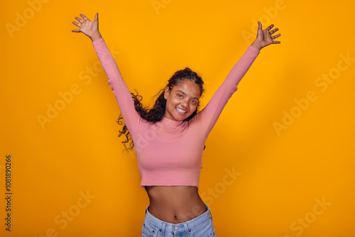 Young Indian woman beams with happiness, her arms stretched wide in celebration. She wears a fitted pink top and casual jeans, radiating positivity against a bright yellow background.