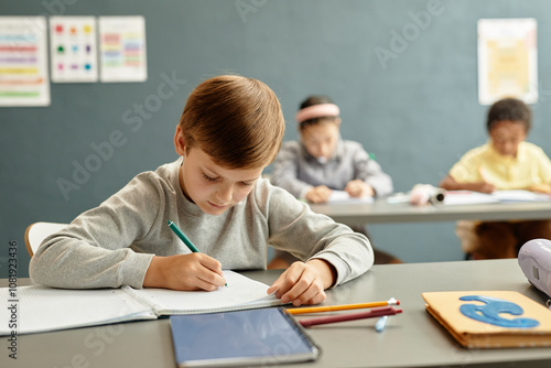 Medium shot of young boy focused on writing task in copybook sitting at desk while studying in classroom with blue wall at school, copy space photo