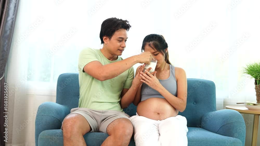 Asian husband takes care of his seven-month pregnant wife by giving her milk from a glass for healthy nutrition and calcium. Pregnant women sit together on the sofa in the living room at home. Asian husband takes care of his seven-month pregnant wife by giving her milk from a glass for healthy nutrition and calcium. Pregnant women sit together on the sofa in the living room at home.