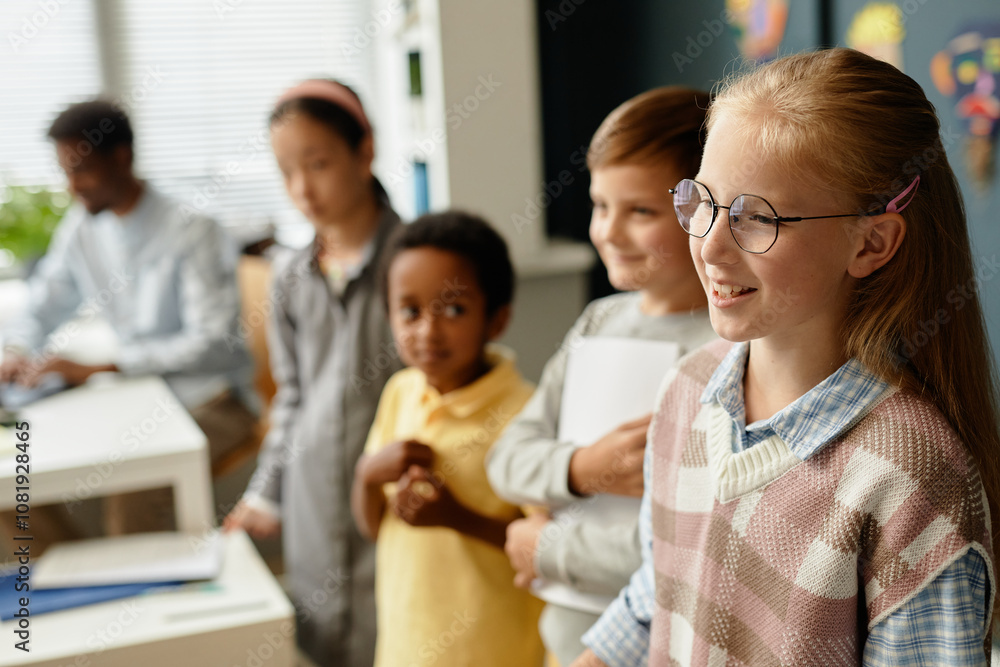 Side view of smiling young girl wearing glasses standing in line with classmates talking in front of class during lesson in primary school classroom, copy space Side view of smiling young girl wearing glasses standing in line with classmates talking in front of class during lesson in primary school classroom, copy space