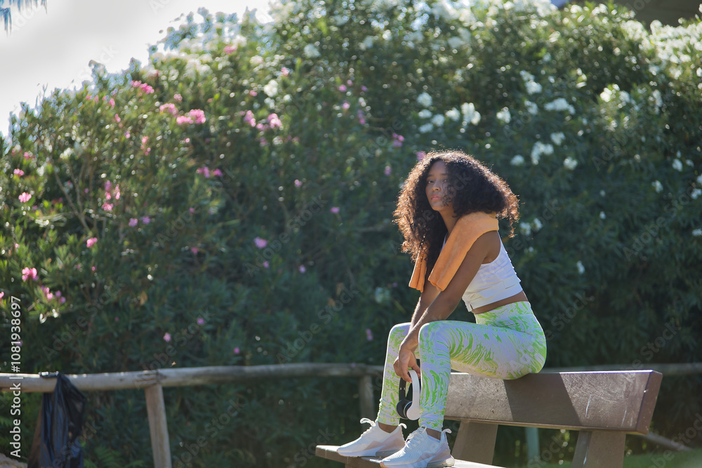 Naklejka premium Latina woman, young, beautiful, brunette, with curly hair, wearing sports clothes, headphones and towel, sitting on a bench relaxed and calm after exercise. Concept sport, exercise, rest, active.